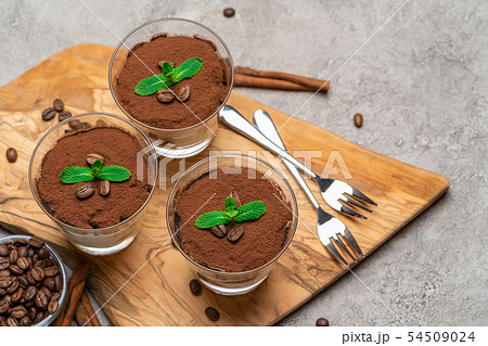 Classic tiramisu dessert in a glass cup on wooden cutting board on concrete background Classic tiramisu dessert in a glass cup on wooden cutting board on concrete background 54509024