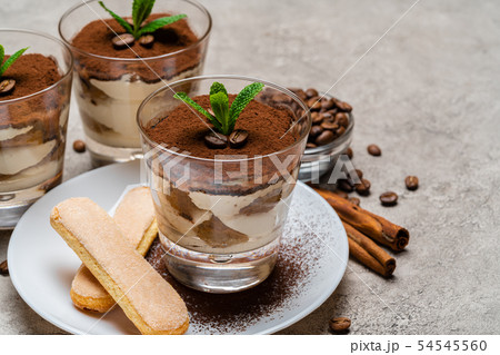 Classic tiramisu dessert in a glass cup and savoiardi cookies on plate on concrete background Classic tiramisu dessert in a glass cup and savoiardi cookies on plate on concrete background 54545560