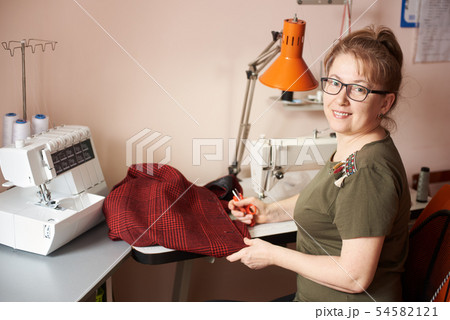 Smiling female tailor on sewing process of skirt, holding scissors. Overlock and sewing-machine on 54582121