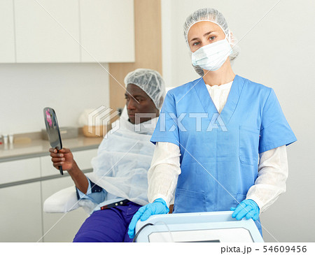 Cosmetician in mask standing before aesthetic facial injection, man with mirror Cosmetician in mask standing before aesthetic facial injection, man with mirror 54609456