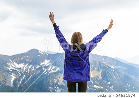 Young blonde girl travelerin a blue jacket raised her hands up and enjoying the green mountain Young blonde girl travelerin a blue jacket raised her hands up and enjoying the green mountain 54621974