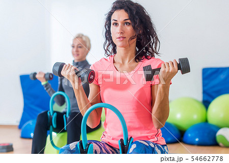 Slim sporty women training sitting on exercise balls holding dumbbells and squeezing Pilates Ring Slim sporty women training sitting on exercise balls holding dumbbells and squeezing Pilates Ring 54662778
