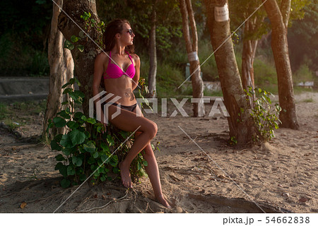 Fitness female brunette model wearing bikini and sunglasses leaning against tree holding her hair 54662838