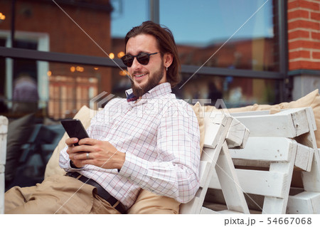 Photo of side of cheerful bearded man in sunglasses with phone in his hands sitting in street cafe Photo of side of cheerful bearded man in sunglasses with phone in his hands sitting in street cafe 54667068