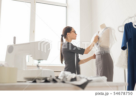 Dressmaker, tailor, fashion and showroom concept - Side view of female fashion designer measuring Dressmaker, tailor, fashion and showroom concept - Side view of female fashion designer measuring 54678750