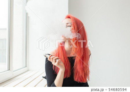 Youth and addiction concept - young red haired woman smoking vape near the window Youth and addiction concept - young red haired woman smoking vape near the window 54680594