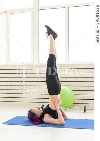 Young slim girl with colored hair makes an headstand on a sports blue mattress next to a barbell and 54681536