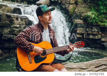 Portrait of a man plays a guitar sitting on a trunk of a tree against a waterfall. Space for your Portrait of a man plays a guitar sitting on a trunk of a tree against a waterfall. Space for your 54702705