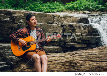 Portrait of a man plays a guitar sitting on a trunk of a tree against a waterfall. Space for your 54702725