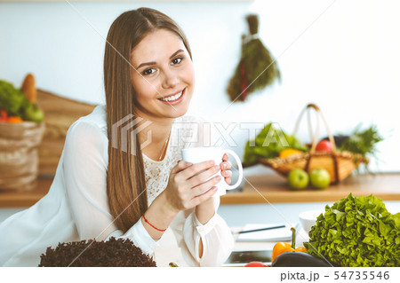 Young happy woman is holding white cup and looking at the camera while sitting at wooden table in Young happy woman is holding white cup and looking at the camera while sitting at wooden table in 54735546