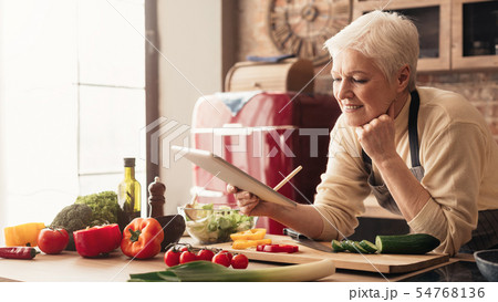 Dreamy senior housewife browsing on digital tablet at kitchen 54768136