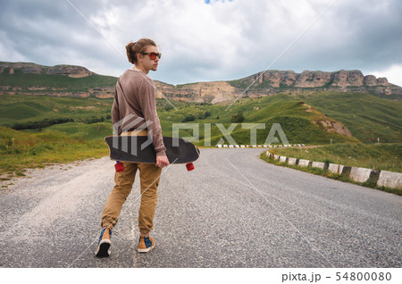 A stylish young man walks along a winding mountain road with a skate or longboard in his hands the 54800080
