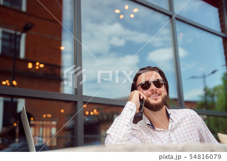Photo of bearded man talking on phone sitting in street cafe in city on summer afternoon 54816079
