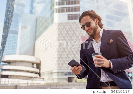 Photo of happy brunet male in sunglasses with phone in his hand while walking through city Photo of happy brunet male in sunglasses with phone in his hand while walking through city 54816117