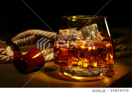 Glass with whiskey, ice cubes and smoking pipe on wooden background. 54854470