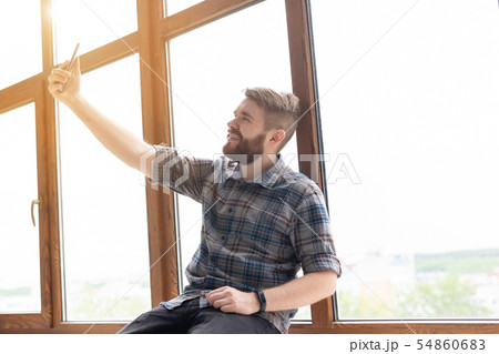Side view of a positive handsome hipster guy and taking a selfie on a smartphone while sitting on a 54860683