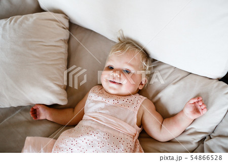 smiling one year old blond girl in a pinkish dress is lying on the sofa at home 54866528