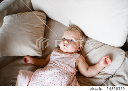 smiling one year old blond girl in a pinkish dress is lying on the sofa at home smiling one year old blond girl in a pinkish dress is lying on the sofa at home 54866529