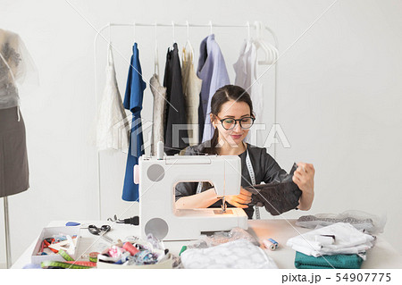 Dressmaker, tailor and fashion concept - Smiling female fashion designer using sewing machine and Dressmaker, tailor and fashion concept - Smiling female fashion designer using sewing machine and 54907775