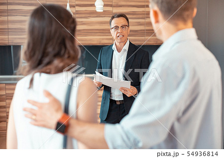 Realtor wearing dark suit holding papers while speaking to clients Realtor wearing dark suit holding papers while speaking to clients 54936814