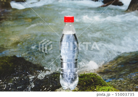 A plastic bottle with a red cap with fresh drinking water against a background of green forest and a 55003735