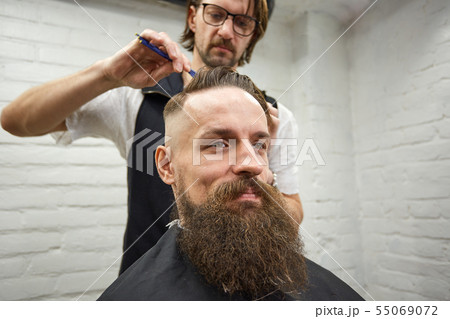Master cuts hair and beard of men in the barbershop, hairdresser makes hairstyle for a young man Master cuts hair and beard of men in the barbershop, hairdresser makes hairstyle for a young man 55069072
