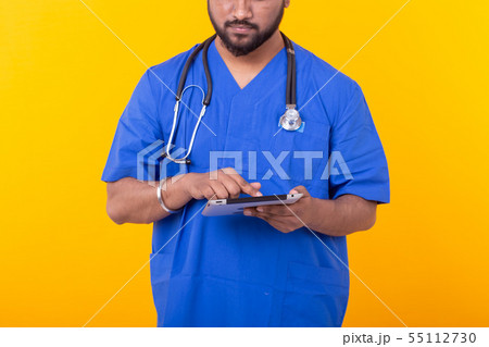 Cute Indian doctor in a blue robe with a stethoscope looking at a tablet based on a yellow Cute Indian doctor in a blue robe with a stethoscope looking at a tablet based on a yellow 55112730