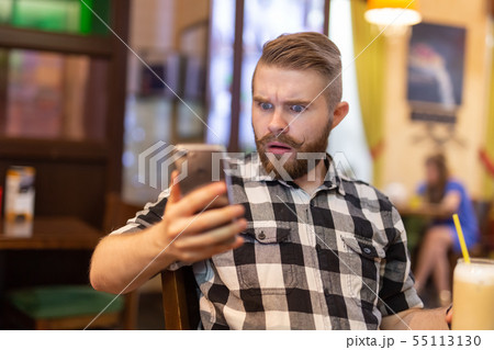 Shocked young hipster man drinking cocoa from a straw and looking at messages in social networks 55113130
