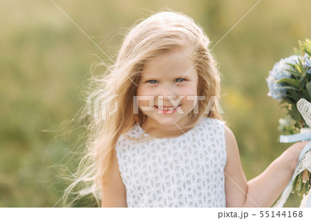 Portrait of beautiful little blond hair girl in field. Girl stand in front of big tree and hold 55144168