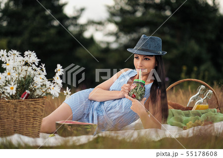 A young beautiful woman in hat and dress is drinking lemonade from a glass jar, sitting on plaid on 55178608