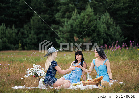 Three young women, blond, brunette and with dyed hair in blue dresses, and hats, sit on plaid and 55178659
