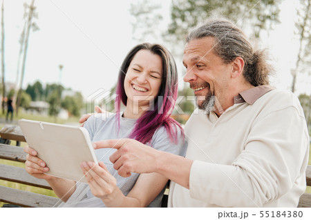 An elderly handsome man with long hair in the tail and a young beautiful woman are sitting on a park 55184380