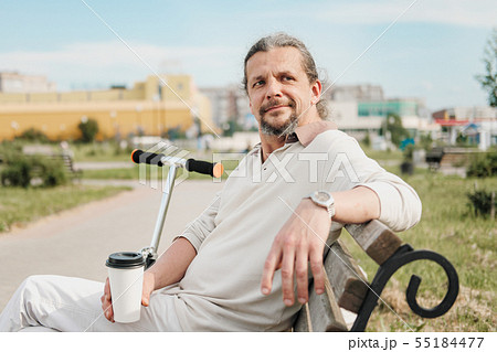 An attractive elderly 50-year-old man with long hair in the tail sits in a public garden on a bench 55184477