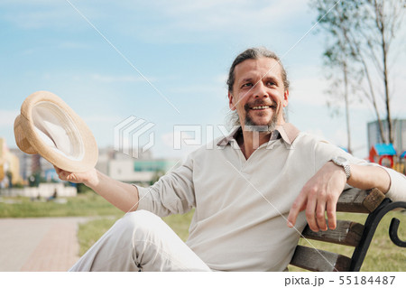 An attractive elderly 50-year-old man with long hair in the tail sits in a public garden on a bench An attractive elderly 50-year-old man with long hair in the tail sits in a public garden on a bench 55184487