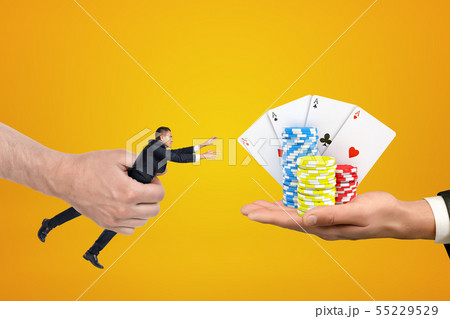 Man's hand holding tiny businessman who is reaching out with both hands for cards and chips on 55229529