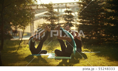 Two young athletic women doing yoga exercises with a trainer in the park surrounded by bright green Two young athletic women doing yoga exercises with a trainer in the park surrounded by bright green 55248333