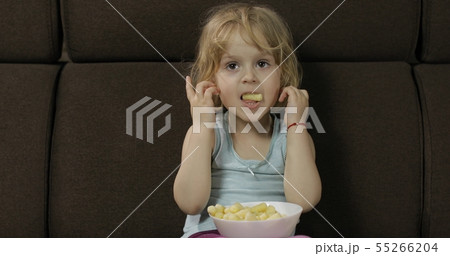 Girl sitting on sofa and eating corn puffs. Child taste puffcorns Girl sitting on sofa and eating corn puffs. Child taste puffcorns 55266204