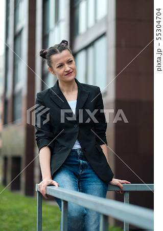 Young attractive woman in black jacket and blue jeans posing outdoor against background of building. 55304704