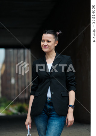Young attractive woman in black jacket and blue jeans posing outdoor against background of building. 55314000