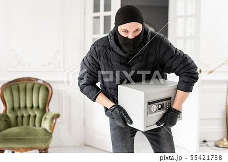 Thief looking at the camera with black balaclava stealing modern Electronic safe box. The burglar Thief looking at the camera with black balaclava stealing modern Electronic safe box. The burglar 55421738