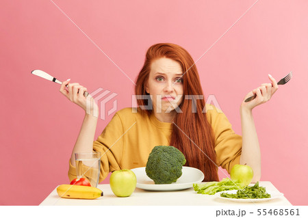 Puzzled, uncertain young red-haired woman sitting at table, served with broccoli Puzzled, uncertain young red-haired woman sitting at table, served with broccoli 55468561