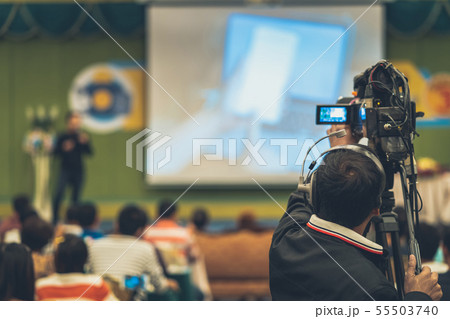 Rear side of Video Cameraman taking photograph to Asian Speaker with casual suit on the stage present the screen in the conference hall or seminar meeting, event and seminar production concept Rear side of Video Cameraman taking photograph to Asian Speaker with casual suit on the stage present the screen in the conference hall or seminar meeting, event and seminar production concept 55503740