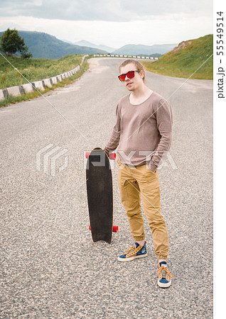 A stylish young man standing along a winding mountain road with a skate or longboard in his hands 55549514
