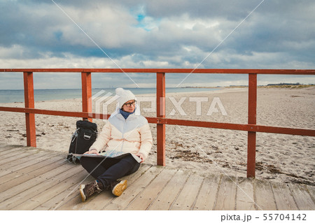 Caucasian woman in hat and jacket with backpack in winter sits on wooden pier on beach near North Caucasian woman in hat and jacket with backpack in winter sits on wooden pier on beach near North 55704142