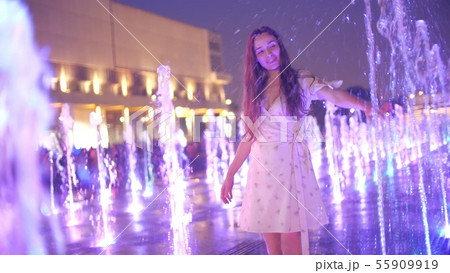 Slim young woman in dress dances in the illuminated fountain in the evening 55909919