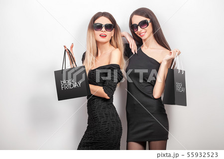 Black friday. Two women wearing black dress and sunglass, with shopping paper bag, look at camera in 55932523