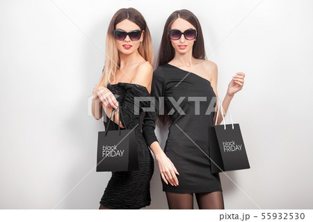 Black friday. Two women wearing black dress and sunglass, with shopping paper bag, look at camera in Black friday. Two women wearing black dress and sunglass, with shopping paper bag, look at camera in 55932530