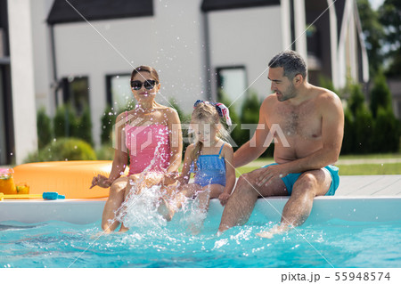 Daughter splashing water while sitting near pool with parents 55948574