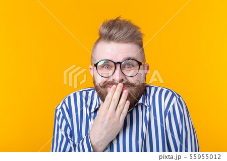 Surprised young male hipster with glasses embarrassed covers his mouth and laughs posing on a yellow 55955012