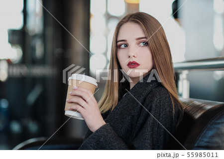 A young beautiful brunette girl in a gray coat is sitting on a bench waiting. Drinking coffee and A young beautiful brunette girl in a gray coat is sitting on a bench waiting. Drinking coffee and 55985011